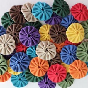 May include: A collection of colorful fabric rosettes, also known as fabric flowers, in various shades of blue, green, red, brown, yellow, and beige. The rosettes are arranged in a circular pattern on a white background.