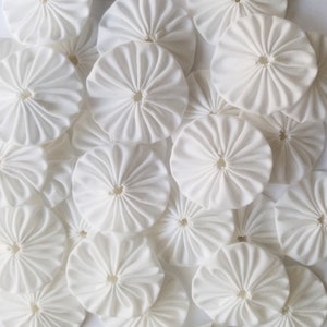 May include: A collection of white fabric rosettes, each with a central hole and a ruffled, pleated design. The rosettes are arranged in a random pattern, creating a textured and visually appealing display.