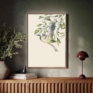 May include: A framed watercolor print featuring a crane in flight, with green and blue plumage, set against a cream background. Green leaves and branches are in the upper right corner. The artwork is displayed on a wooden cabinet.
