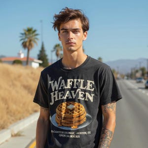 May include: Black t-shirt with the words "Waffle Heaven" in a distressed font. Below the text is an illustration of a stack of waffles with butter and syrup. The shirt also includes the address "Carter US Hwy, Austin, TX" and "Open 24 Hours."