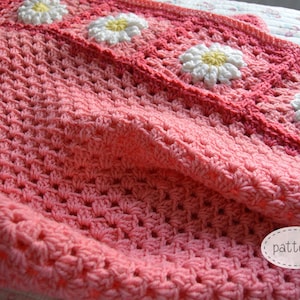 May include: A pink and white crochet baby blanket with a daisy pattern. The blanket is folded in half and has a "pattern" label on the bottom right corner.