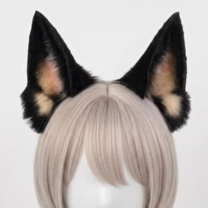 Könnte beinhalten: Ein Paar spitz zulaufende Tierohren an einem Haarreif, mit schwarzem Kunstfell an den Außenkanten und einem weichen, hellbraunen und rosafarbenen Innenbereich. Die Ohren sind auf einem weißen Schaufensterpuppenkopf mit hellem Haar platziert.