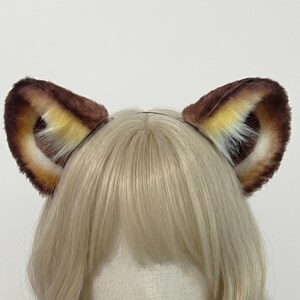 Puede incluir: Una diadema con un par de orejas de gato de piel sintética. Las orejas son marrones con detalles en beige y blanco, creando una apariencia esponjosa. La diadema es de metal fino. El accesorio está diseñado para ser usado en la cabeza.