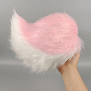 Può includere: Un accessorio a forma di coda di animale in pelliccia sintetica, caratterizzato da un design bicolore con pelo rosa pastello morbido nella parte superiore e pelo bianco brillante nella parte inferiore, con una forma curva e affusolata e fibre lunghe.