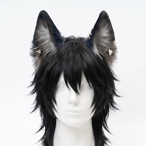 Puede incluir: Un par de orejas de animal de piel sintética en tonos negro, gris y blanco, con tachuelas piramidales plateadas y pequeños aros sujetos a los lados. Las orejas están colocadas sobre una peluca sintética negra, escalonada.