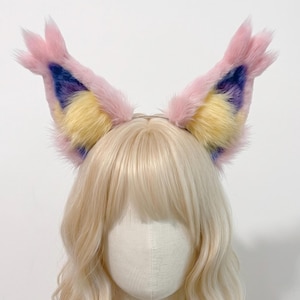 May include: A pair of tall, pointed animal ears attached to a thin headband. The ears are covered in long, soft faux fur featuring a color palette of pastel pink on the outer edges, bright yellow in the center, and deep purple at the base.