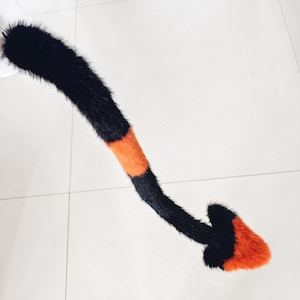 Puede incluir: Un accesorio de cola de piel sintética largo y flexible con un patrón de rayas en secciones negras y naranja brillante. La cola termina en una punta distintiva en forma de corazón de color naranja vibrante, creando un contraste con el pelaje.
