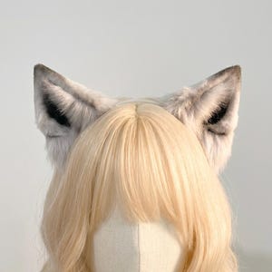 Könnte beinhalten: Ein Paar graue und schwarze Wolfsohren aus Kunstfell auf einem Stirnband. Die Ohren sind über einem Schaufensterkopf mit blonden Haaren und einer weißen Gesichtsbedeckung positioniert. Die Ohren haben ein realistisches Aussehen.