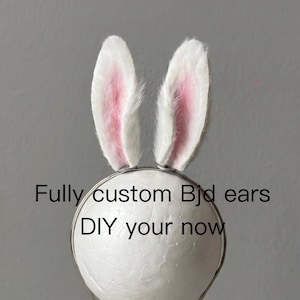 May include: A pair of white and pink faux fur rabbit ears attached to a thin metal headband. The ears are upright and textured with soft white fur on the outer edges and pink fur in the center. The text "Fully custom Bjd ears DIY your now" is visible.