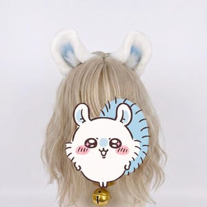 May include: A headband featuring two plush white animal ears with soft blue inner coloring, worn on a blonde wig. A gold-toned metal bell hangs from the base of the wig, and a graphic illustration of a white creature with blue accents is overlaid.