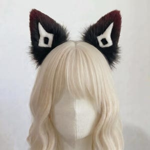 Può includere: Un cerchietto con due orecchie da animale a punta in pelliccia sintetica. Le orecchie sono principalmente nere con punte rosso scuro e presentano un motivo geometrico bianco a forma di rombo sulla parte anteriore, esposto su un manichino.