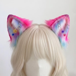 Puede incluir: Un par de orejas de gato en una diadema de piel sintética con un degradado en rosa, azul y blanco. Las orejas están decoradas con dijes de cruz plateados, pequeños imperdibles y detalles en forma de estrella de colores. Las puntas tienen un acabado brillante.