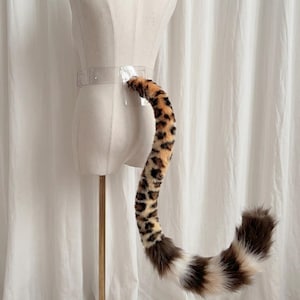 May include: A faux fur tail with a leopard print pattern, attached to a clear belt. The tail is brown, tan, and black, and is displayed on a mannequin. The tail is approximately 76 cm long.