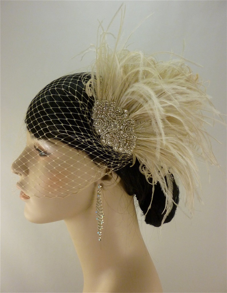 May include: A white feather headpiece with a crystal embellishment and a birdcage veil. The headpiece is worn on a mannequin with long, dark hair.