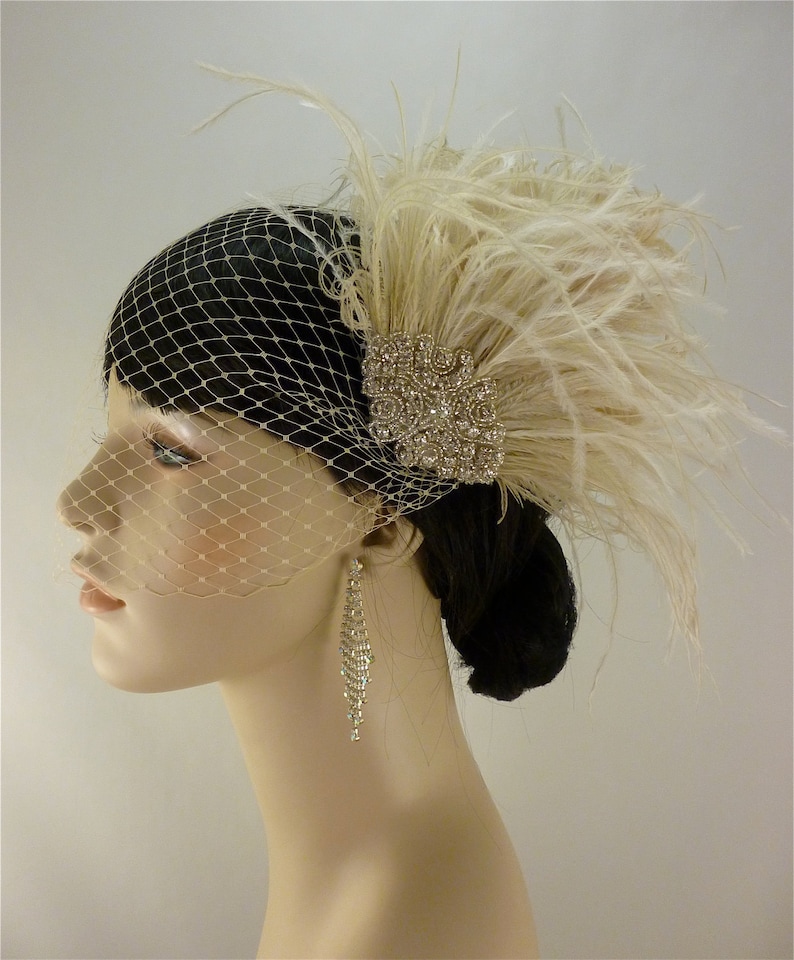 May include: A white feather bridal hairpiece with a crystal embellishment and a birdcage veil. The hairpiece is worn on a mannequin with long, dark hair.