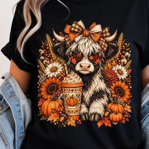 May include: A black t-shirt featuring a graphic of a fluffy highland cow wearing a plaid bow and heart-shaped sunglasses. The cow is holding a coffee cup surrounded by sunflowers, pumpkins, and autumn leaves. The text ElvoraStudioMA is visible.