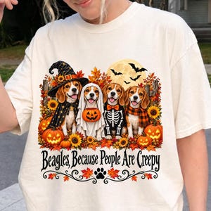 Op de afbeelding: Een crèmekleurig t-shirt met een afbeelding van vier Beagle-honden in Halloween-kostuums, waaronder een heks, een geest, een skelet en een hond met een sjaal, omringd door pompoenen, zonnebloemen en vleermuizen, met de tekst Beagles, Because People Are Creepy.