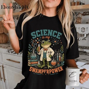 May include: Black t-shirt with a colourful graphic of a frog in a lab coat holding a test tube. The text "SCIENCE is my SWAMPFRPOWER" is displayed above and below the frog illustration. A mug with the letter "E" is also visible.