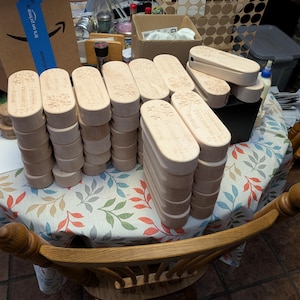 May include: Several stacks of unfinished wooden boxes with oval lids are displayed on a floral tablecloth. The lids are engraved with text and a floral design. The boxes are cylindrical with a flat base. The boxes are made of light-coloured wood.