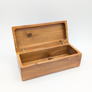 Peut inclure: Boîte en bois ouverte avec un couvercle à charnière. La boîte est en bois brun clair avec un grain visible et un nœud sur le couvercle. L'intérieur est vide et la boîte est posée sur un fond blanc.