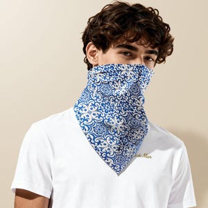 Peut inclure: Un t-shirt blanc avec le texte "do Mar" en lettres dorées. La personne porte un bandana bleu et blanc à motifs sur le visage. Le bandana présente un motif floral.