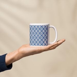 Peut inclure: Mug en céramique blanche avec un motif géométrique bleu et blanc. La tasse a une anse incurvée et une forme cylindrique. Le motif est un motif répétitif de formes florales stylisées. La tasse est tenue dans une main.