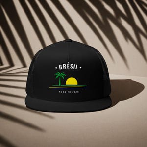 Peut inclure: Casquette de camionneur noire avec un dos en filet et une visière plate. Le devant affiche le mot "Brésil" au-dessus d'un palmier et d'un soleil jaune. Le texte "Road to 2026" est en dessous.