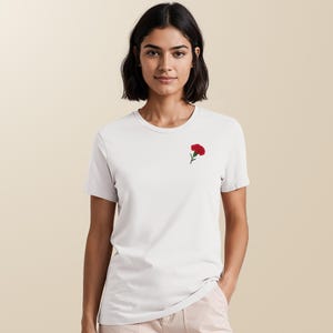 May include: A white t-shirt with a small, embroidered red carnation flower on the left chest. The t-shirt has short sleeves and a crew neck. The model is wearing light pink pants.
