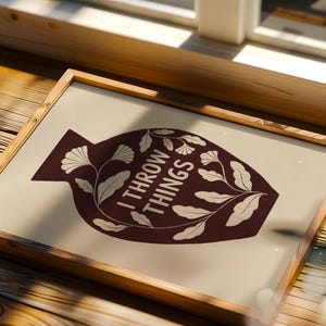 May include: A framed print featuring a dark maroon silhouette of a vase shape on a light beige background. The vase contains the white text I THROW THINGS surrounded by white botanical illustrations of leaves and flowers on thin stems.