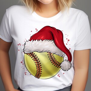 May include: White t-shirt featuring a yellow softball with red stitching, wearing a red Santa hat with white trim and a pom-pom. The design includes red splatter accents, creating a festive holiday theme.