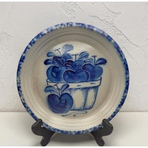 May include: A ceramic plate with a blue and white apple basket design. The plate has a cream-colored center with a blue border. The apple basket is in the center of the plate and is painted in blue.