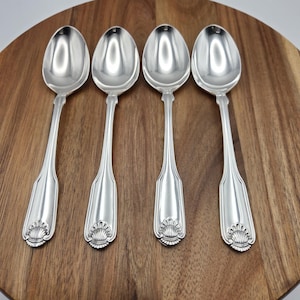 May include: Four silver spoons with an oval bowl and ornate handle, featuring a shell design at the base. The spoons are arranged on a wooden surface, reflecting light. A classic silverware set.