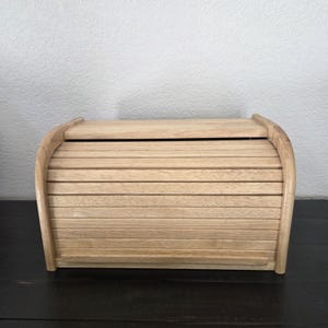 May include: A light brown wooden bread box featuring a curved roll-top lid with horizontal slats. The storage container has a smooth finish and a rectangular base, designed with a sliding door mechanism to access the interior compartment.