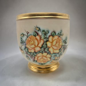 May include: A cream-colored porcelain cup featuring a hand-painted floral design of three large yellow-orange roses with green leaves and stems. The cup has a rounded body, a flared base, and a gold-painted rim and base edge for a polished finish.