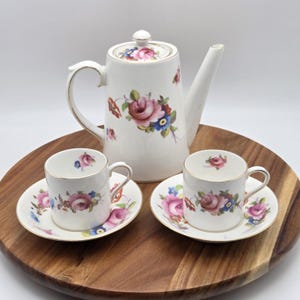 May include: A white porcelain tea set with a floral rose pattern. The set includes a teapot, two teacups, and saucers. The cups and saucers have gold trim. The teapot is approximately 9 inches tall.