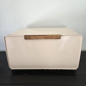 May include: A rectangular, cream-colored bread box with a smooth, glossy finish. The front panel features a horizontal, textured wooden handle centered near the top edge. The container has rounded corners and a clean, solid-colored exterior design.