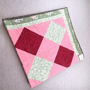 May include: A square quilted fabric piece featuring a patchwork design of solid pink, dark red with swirling stitch patterns, and light green squares with a small floral print. The border is a green fabric with a blue and pink floral pattern.