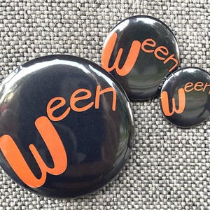 May include: Three black, circular badges with the word "Ween" in orange lettering. The badges are of varying sizes and are arranged on a textured grey surface. These are likely Halloween-themed.