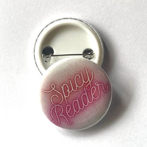 May include: A round, white button with the text "Spicy Reader" in a pink, cursive font. The button has a pin on the back and a silver metal rim. The background is white.