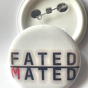 May include: A white button with the words "FATED" and "MATED" printed in black, with the "M" in "MATED" in red. The button is round and has a pin back.
