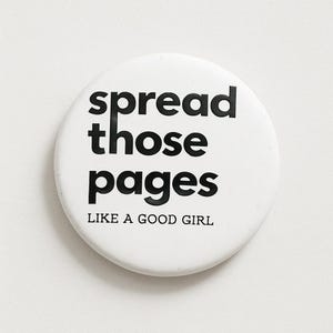 Op de afbeelding: Een witte knoop met zwarte tekst: "spread those pages LIKE A GOOD GIRL". De knoop is rond en heeft een eenvoudig ontwerp. De tekst is in een vetgedrukte, schreefloze lettertype.