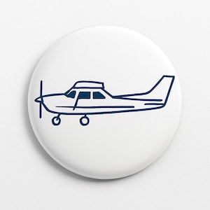 May include: A white button featuring a navy blue outline of a small aircraft. The plane has a propeller, windows, and a tail. The button is circular with a smooth, glossy finish.