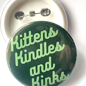 May include: A round, dark green button with the words "Kittens Kindles and Kinks" in light green script. The button has a pin back and a white plastic backing. The button is a fun accessory.
