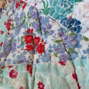 May include: Close-up of a patchwork quilt with a floral pattern. The quilt features a variety of colors, including red, blue, purple, and green. The fabric appears to be cotton, and the quilt is quilted with a diamond pattern.