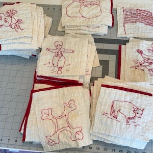 May include: A collection of square white fabric quilt blocks featuring red embroidered designs including a flower, a child, an American flag, a bird, a geometric shape, and a cow. Each block has a red stitched border on one edge.