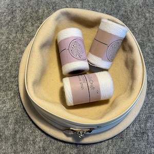 May include: A cream-colored hat with a decorative band holds three spools of white thread. Each spool has a pink label with a floral design and text. The hat sits on a gray surface.