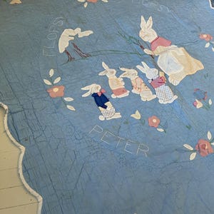 May include: A light blue quilt with a scalloped edge, featuring appliqued rabbits and floral designs. The quilt has the names "Peter", "Flopsy", and "Cottontail" embroidered on it. The quilt is a vintage, handmade item.