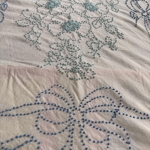 May include: A white fabric textile featuring intricate blue embroidery. The design includes a central floral motif composed of small dots and lines, accompanied by a prominent bow pattern at the bottom, all rendered in a consistent blue thread.