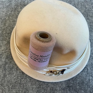 May include: A cream-colored cloche hat with a white ribbon and a decorative jeweled accent. A spool of pink thread with the text "Creative Needle" is on the hat. The hat is on a gray surface.