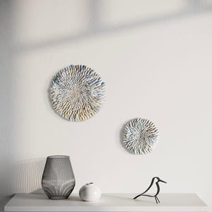 Könnte beinhalten: Zwei kreisförmige Wandkunstwerke mit strukturierten, geschichteten Designs in Weiß-, Blau- und Beigetönen. Eine schwarze Metallvase, eine kleine weiße Vase und eine schwarze Metallvogelskulptur stehen auf einem weißen Regal.